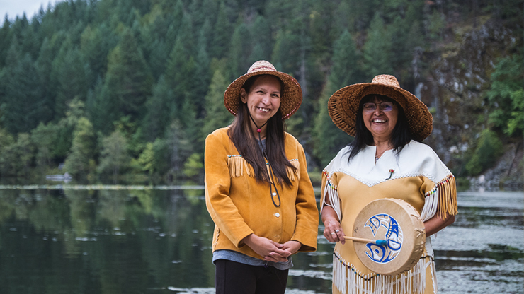  Alaska Native women from two generations stand together lakeside. One holds a Tribal drum, while the other holds her pregnant stomach. Both wearing traditional clothing, and headwear.
