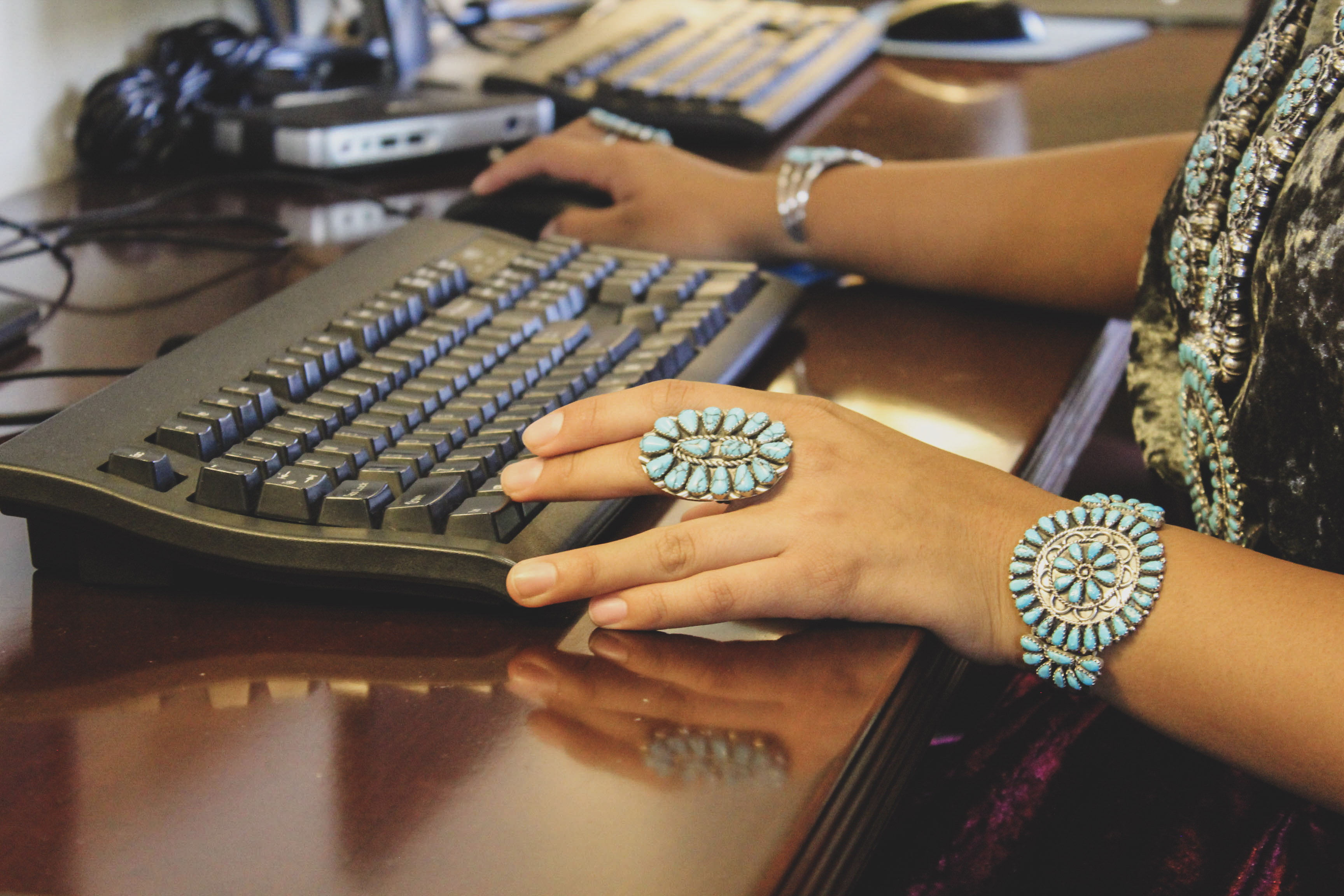 Navajo youth works on computer in computer lab. Two hands using a computer mouse and keyboard. Person wearing traditional Navajo turquoise and silver rings, bracelets and necklace.
