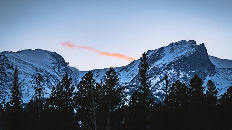 Mountain landscape. Mountain covered in snow. With trees at the base. Small line of clouds are bright orange. 