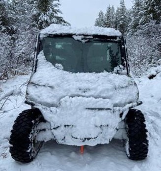 A snow-covered utility side-by-side vehicle sits within a snowy forest trail.