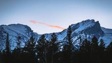 Mountain landscape. Mountain covered in snow. With trees at the base. Small line of clouds are bright orange. 