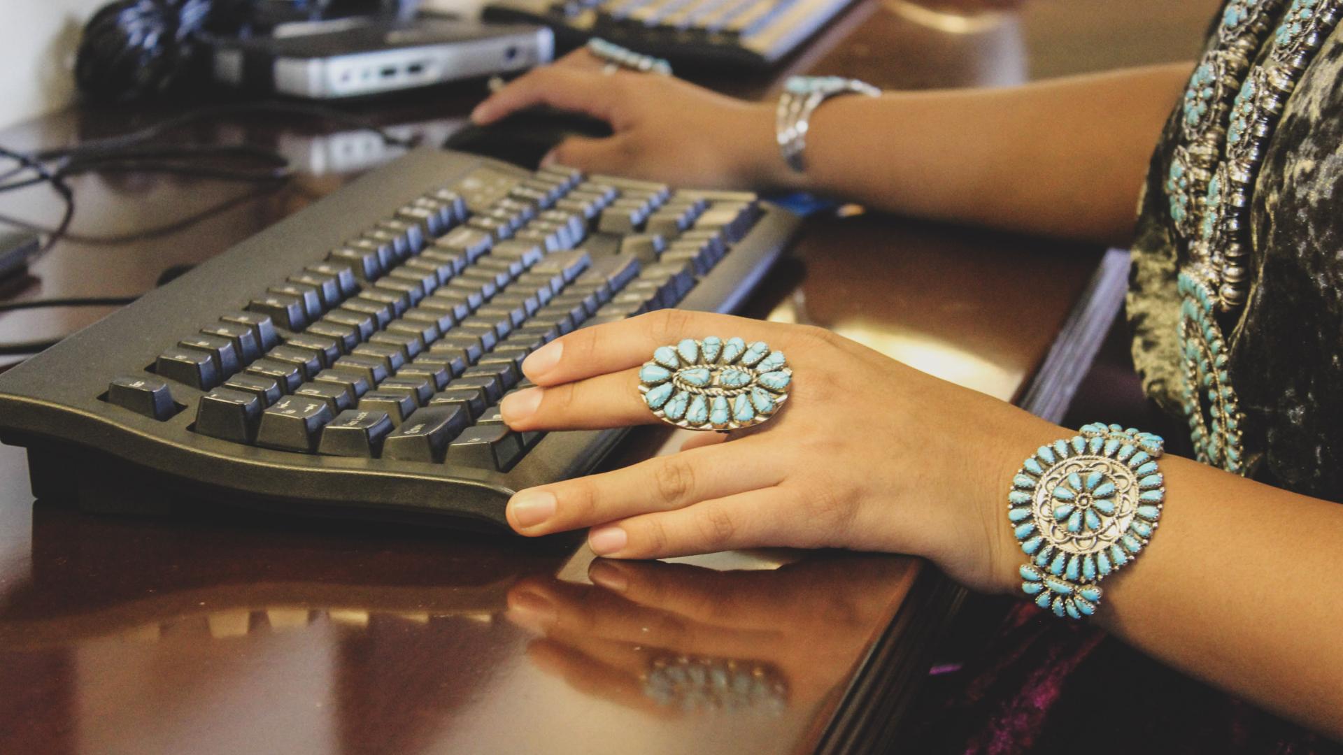 Two hands using a computer mouse and keyboard. Person wearing traditional Navajo turquoise and silver rings, bracelets and necklace. 