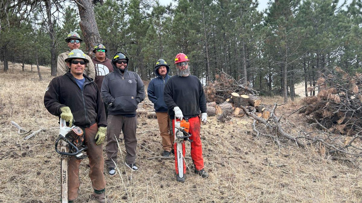 6 wildland firefighters from the BIA Pine Ridge Agency stand outside in front of a tree line on the Oglala Sioux Reservation.