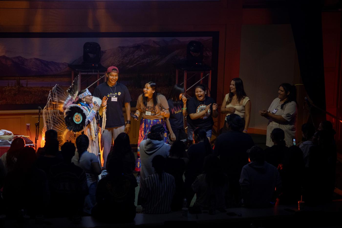 On stage, an Indigenous person dressed in regalia speaks into a microphone while Indigenous youth laugh and clap in front of a crowd.