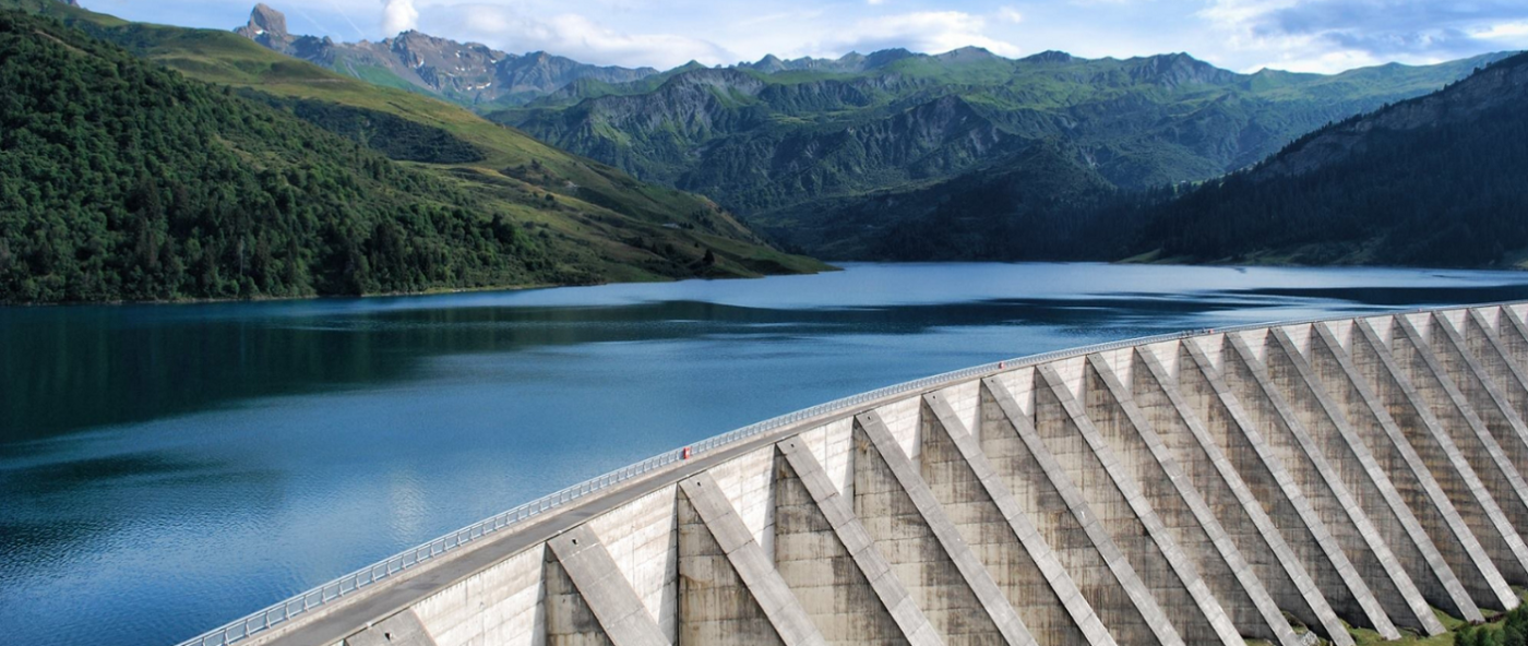 The repeating lines in of a white dam structure contrasts with the natural beauty of a placid blue body of water surrounded by lush green hills.