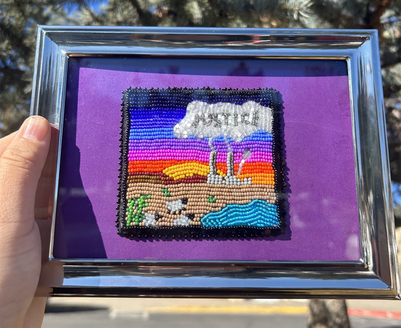 Beadwork landscape of a sunset with sheep grazing by a pool of water in the foreground. In the background an industrial building with tall smokestacks emits a plume of smoke which reads "INJUSTICE."