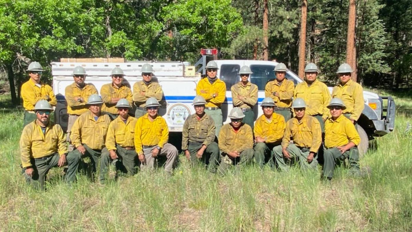 A photo taken in 2021 of the Navajo Hotshots crew in uniform, kneeling outside in the grass in front of a fire vehicle.