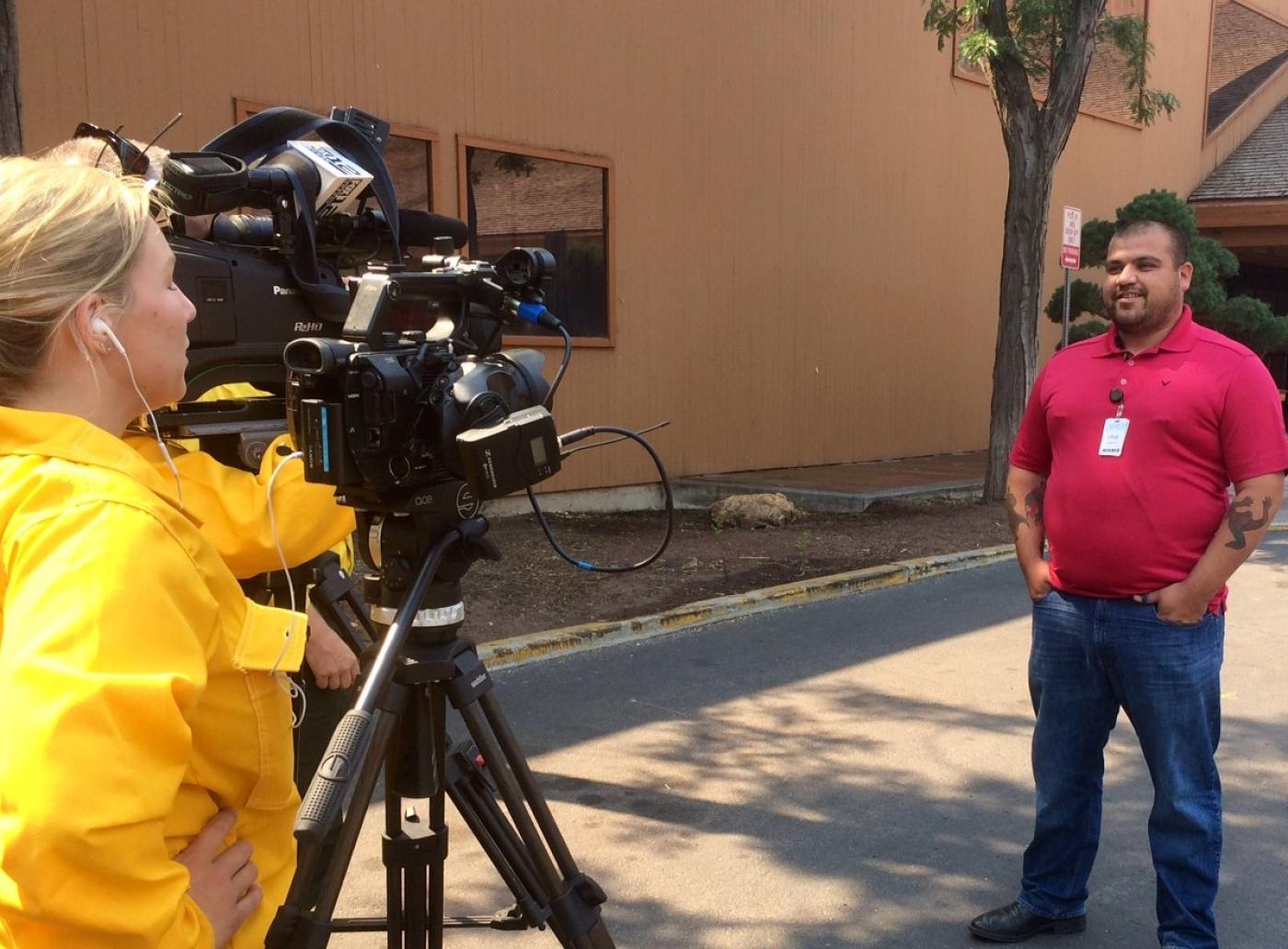 A public information officer records an on-camera interview with an individual on an empty street.