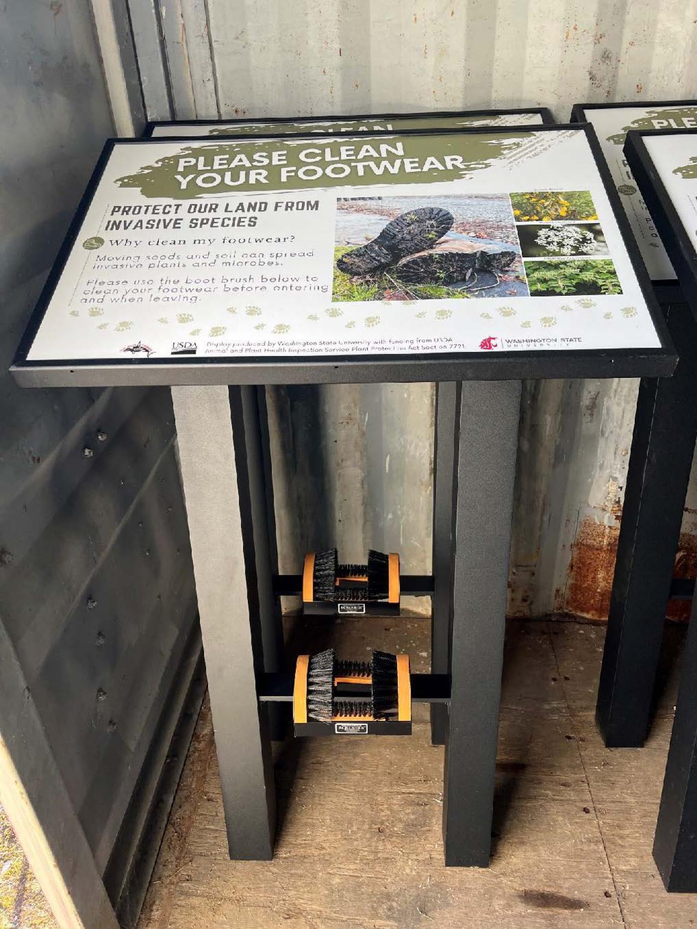 A boot brush station with a sign explaining how cleaning footwear helps prevent the spread of invasive plant species. The station has two mounted brushes for scrubbing shoes and is set inside a metal-sided structure.